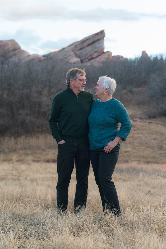 An older couple in sweaters smiling at each other in a dry grass field below red rock formations