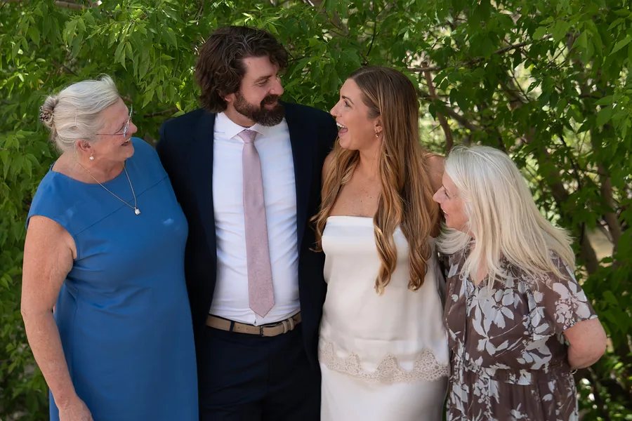 A newly married couple laughing with both of their moms under a green tree