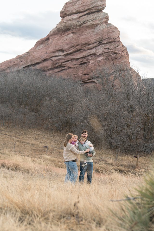 A mom and dad each holding a small child in tall golden grass beneath a towering red rock