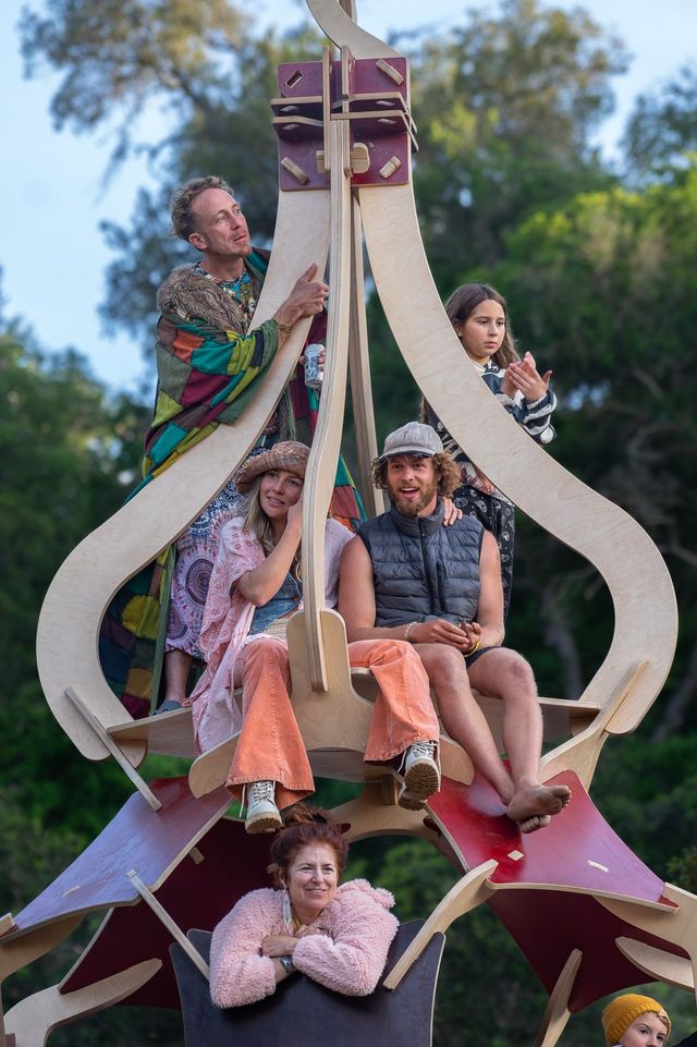 Friends and family of all ages perched together on a big wooden art installation among the trees