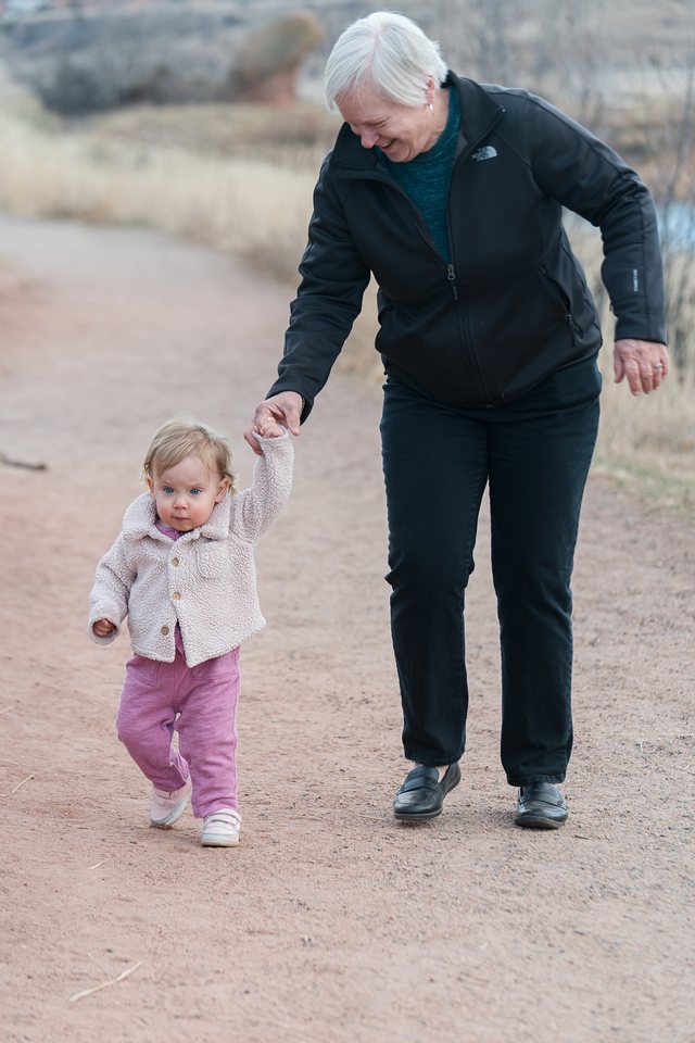 A grandma laughing as she holds her toddler granddaughter's hand on a dirt trail