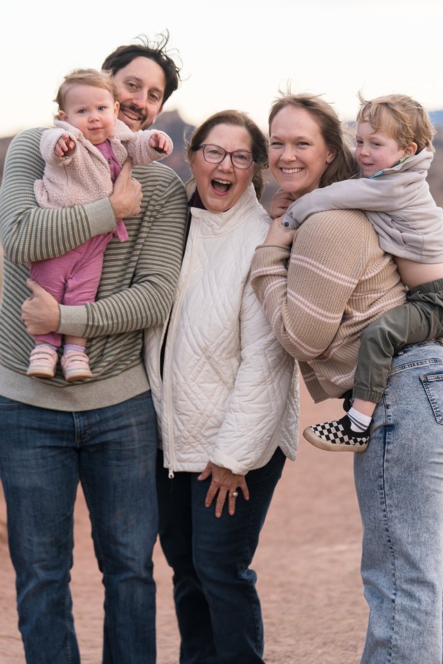 A grandma cracking up between her grown kids, one holding a pointing baby and the other giving a toddler a piggyback