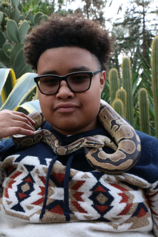 A teen in glasses with his ball python draped around his shoulders in a cactus garden