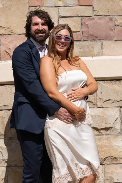 A joyful couple embracing in front of a stone wall, the bride in a white dress