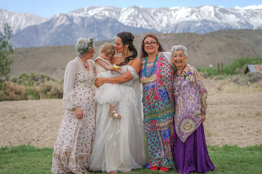Three generations of women in flowing, colorful dresses standing together in a field below snow-capped mountains