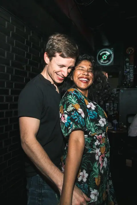 A couple laughing with their eyes closed, dancing close against a dark brick wall