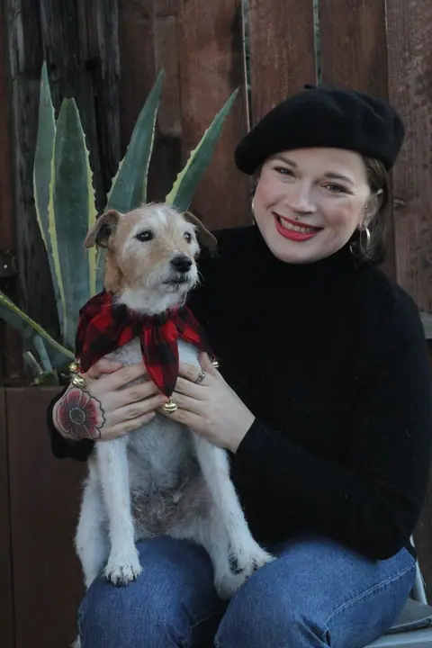 A woman in a black beret and turtleneck holding a small terrier in a plaid scarf